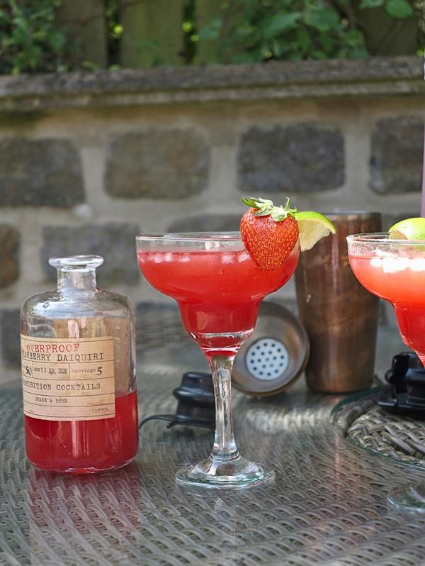 A clear glass bottle labeled 'Overproof Strawberry Daiquri' from Prohibition Cocktails filled with red liquid sits on a metal patio table beside two pink cocktails garnished with strawberries and lime wedges, with a stone wall and greenery in the background.