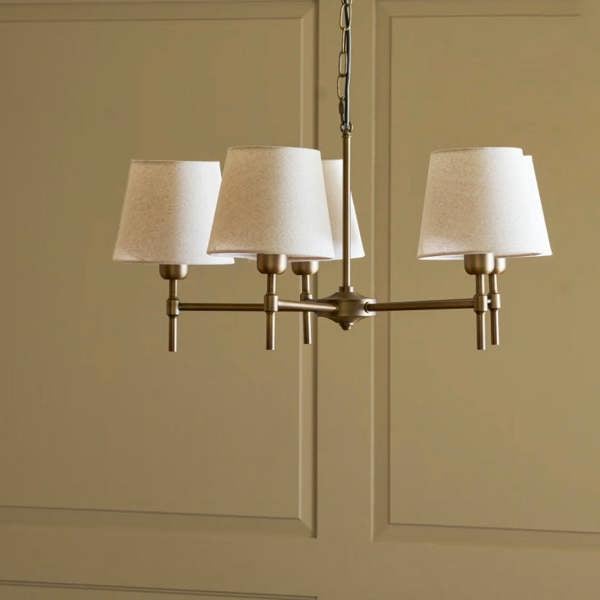 A 5-light antique bronze steel chandelier with cream linen shades hanging against a olive-green panelled wall by Abode Home & Gifts.