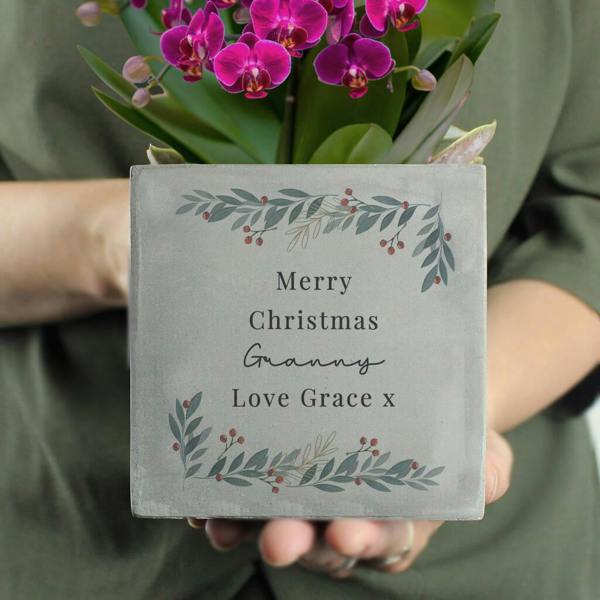 A pair of hands holding a square, grey concrete planter featuring a festive wreath illustration with the text 'Merry Christmas Granny Love Grace x' and a vibrant purple orchid plant in the background.