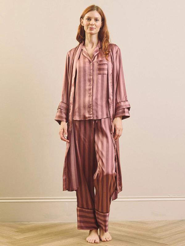 A woman modeling a matching two-piece pajama set with a long-sleeve button-front top and wide-leg pants and matching robe, both in a dusty rose and brown vertical stripe pattern, against a plain beige wall.
