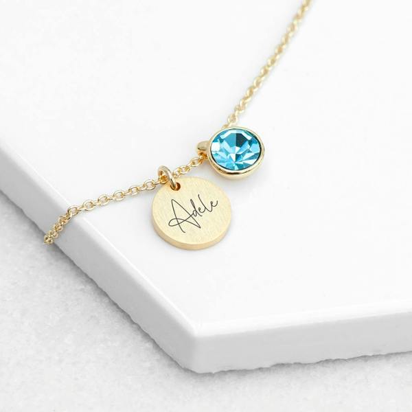 A close-up shot of a delicate gold chain necklace featuring a small, glistening birthstone charm next to a round gold disc, elegantly engraved with the name 'Adele' in a sophisticated script font.