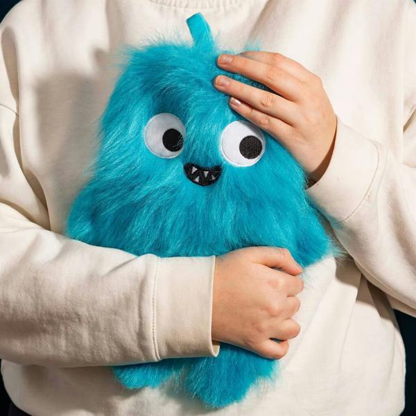 A person wearing a white sweatshirt closely hugging a plush blue monster hot water bottle with shaggy fur and wide cartoon eyes.