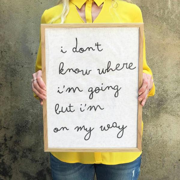 A woman in a bright yellow shirt holding a large solid oak framed print made of white cotton fabric with the lowercase handwritten-style text "i don't know where i'm going but i'm on my way"