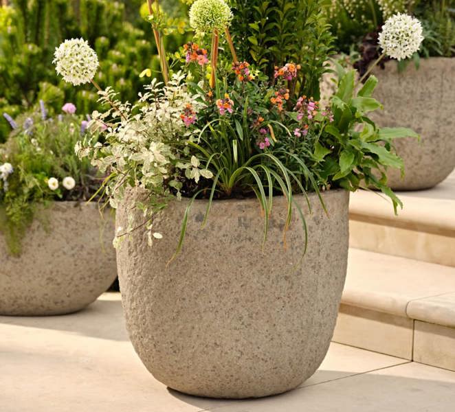 Cranford round garden planters, shown in a rustic, textured ficonstone finish resembling weathered light grey stone. The planters feature varied summer plantings including alliums and ornamental grasses, set upon stone patio steps in a sun-drenched outdoor living area. The aesthetic leans heavily into modern coastal garden styling and al fresco dining decor.