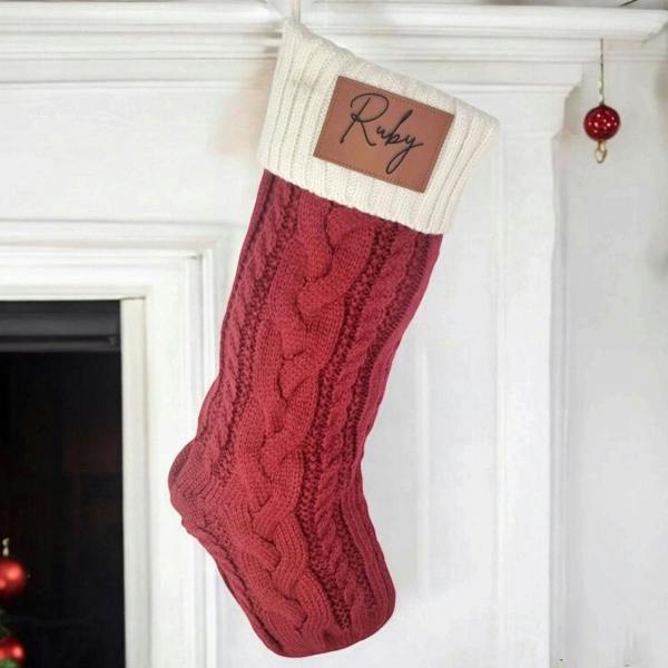 Personalised red cable-knit Christmas stocking with an engraved faux leather patch reading Ruby hanging from a white mantlepiece.