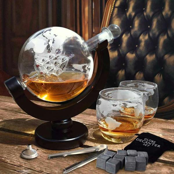 A hand-blown glass globe decanter filled with an amber spirit, featuring a small ship inside and etched world map markings, resting on a dark wooden rotating crescent stand. Two matching etched globe-style glasses sit next to it on a wooden table, alongside whiskey chilling stones and metal tongs.